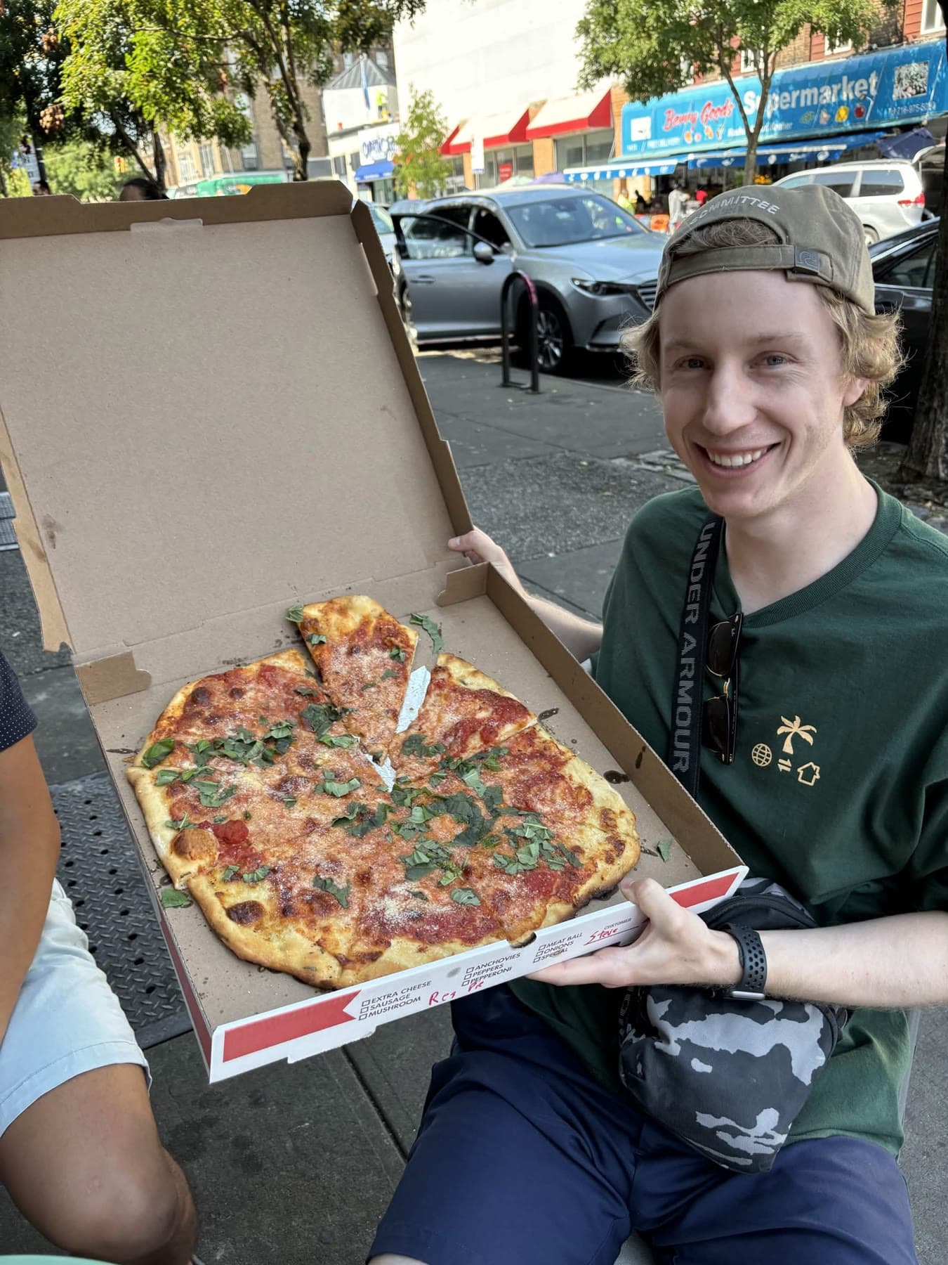 Garrett on a New York sidewalk holding open a box with a whole Di Fara pie topped with basil.