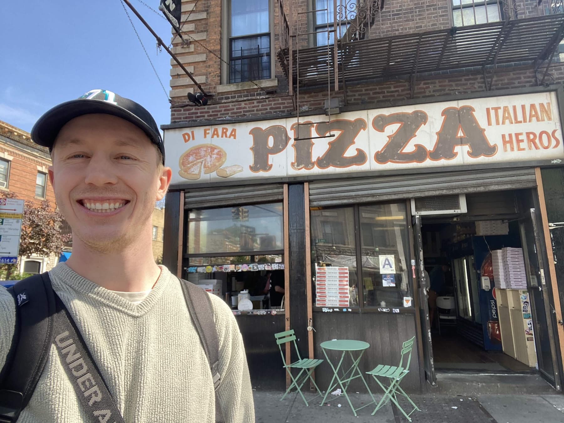 Garrett smiling outside Di Fara Pizza in Brooklyn, under its hand-painted PIZZA sign.